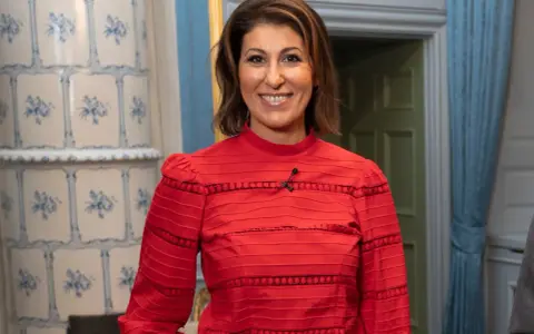 Rania Shemoun Olsson ler i en röd blus med puffärmar. Bakom henne syns en kakelugn och en dörröppning.