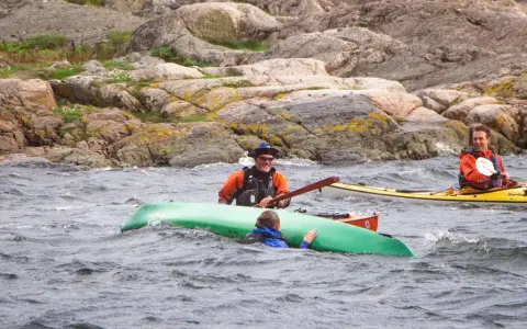 Två män i kajaker nära en klippig kust. En person i en grön kajak verkar ha problem. En diskussion om kanot vs kajak kan uppstå.