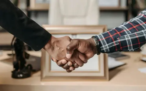 Uścisk dłoni symbolizujący udaną **pracę w Anglii**. Dwie osoby, jedna w czarnej marynarce, druga w kratowanej koszuli, potwierdzają porozumienie.