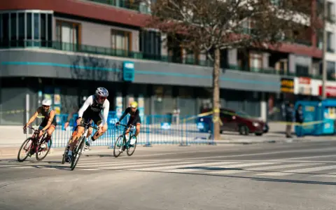 Cyclists race through a city street, perhaps part of le grand tour de France, with buildings and spectators lining the course.
