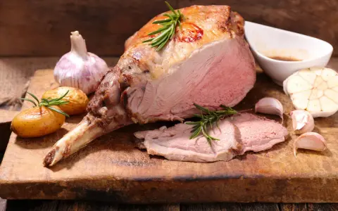 Gigot d'agneau rôti, parfait pour une recette d'agneau pascal. Accompagné de pommes de terre, ail et romarin sur une planche en bois.