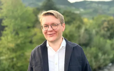 Ein Mann mit Brille lächelt in die Kamera. Er trägt ein weißes Hemd und eine dunkle Jacke. Im Hintergrund sind Bäume und Berge zu sehen. Er studiert Seelsorge.