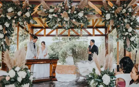 Novios celebran aniversario en altar decorado con flores y pampas. Un oficiante habla mientras la pareja sonríe.
