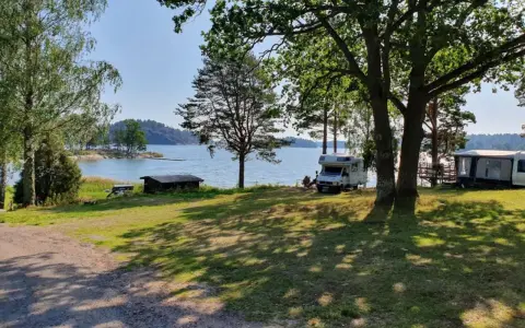 Solig dag vid vattnet på Kalvholmens camping. Husbilar och husvagnar står uppställda på gräsmattan nära sjön.