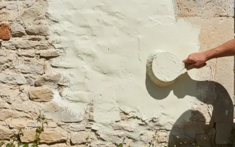 Une main applique une peinture claire sur un mur en pierre enduit de chaux. La question "peut on peindre sur de la chaux" trouve ici sa réponse visuelle.