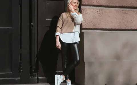 Eine Frau kombiniert elegant schwarze Hose mit einem zweifarbigen Pullover und weißem Hemd. Weiße Plateau-Sneaker runden den Look ab.