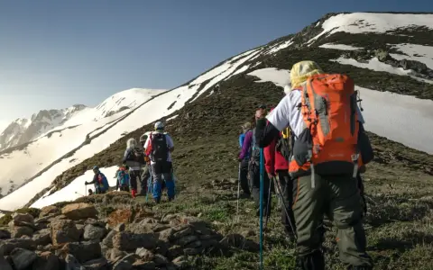 Un groupe de randonneurs progresse sur un sentier rocailleux, le soleil éclairant les montagnes enneigées. Ils semblent déterminés à savoir son altitude.