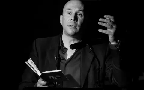 Un uomo calvo, narratore protagonista, legge da un libro aperto, gesticolando con la mano.
