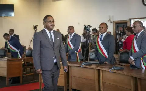 Des hommes en costume, certains portant des écharpes aux couleurs nationales, se tiennent dans une salle. L'un d'eux semble être une persona non grata, marchant à l'écart.