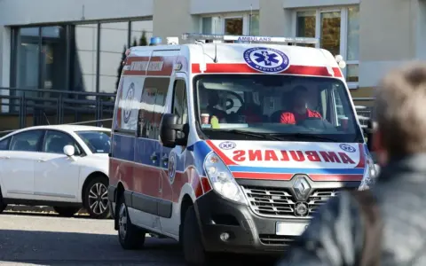 Ambulans z ratownikami medycznymi na tle budynku i białego samochodu.