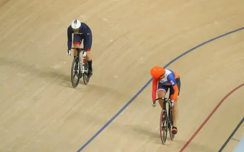 Two cyclists compete in a track sprint. One rider in a blue and white uniform leads, while another in orange and blue follows closely.