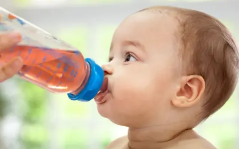 Kleines Baby trinkt aus einer Flasche mit rosafarbenem Tee. Ab wann Tee Baby? Diese Frage stellen sich viele Eltern.