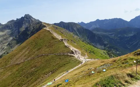 Kamienista ścieżka wijąca się po zielonym zboczu górskim, z turystami i skalistymi szczytami w tle.