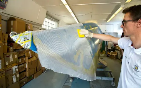 Mann bearbeitet mit gelbem Werkzeug und Handschuh ein Bootsteil. Epoxidharz Bootsbau-Material wird aufgetragen.