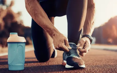 Uomo si allaccia le scarpe da corsa, pronto per correre a stomaco vuoto. Accanto, una borraccia blu.