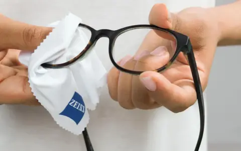 Dłonie czyszczą okulary okularowe białą ściereczką z logo Zeiss.