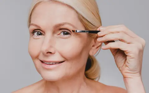 Une femme blonde applique du maquillage sur son œil, son sourcil blanc est visible.