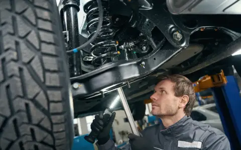 Mechanik bada elementy zawieszenia samochodu, oświetlając je latarką. Widoczna sprężyna i amortyzator.