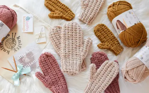 Mitones tejidos a mano con patrón de dos agujas. Varios pares en tonos rosa, beige y mostaza, junto a ovillos de lana y agujas.