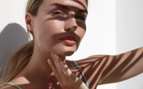 Eine Frau streicht sich sanft über den Hals, wo leichte Falten zu sehen sind. Sonnenlicht wirft Schatten von Palmblättern auf ihre Haut.