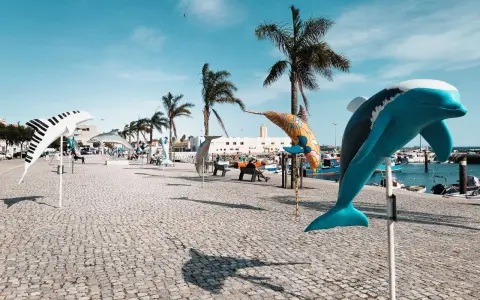 Bunte Delfin-Skulpturen säumen die Promenade in Setúbal, Portugal, unter blauem Himmel und Palmen.