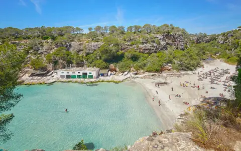 Türkisfarbenes Wasser und ein sonniger Sandstrand in Cala Pi, umgeben von grünen Hügeln. Menschen entspannen sich unter Sonnenschirmen.