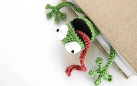 Marcador de libro con ranas amigurumi, verde y negro, con lengua roja, asomando de un libro beige.