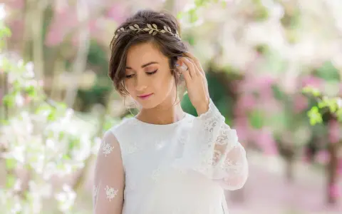 Delikatna tiara na głowę zdobi brunetkę w białej sukni z koronkowymi rękawami, otoczoną kwitnącymi drzewami.