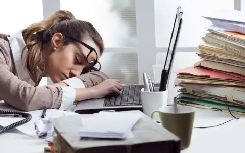Zmęczona kobieta śpi na laptopie, otoczona stertami dokumentów. To obraz pracoholizmu, który doprowadził ją do wyczerpania.