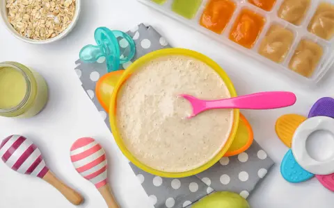 Un tazón de papilla de cereales con una cuchara rosa, junto a avena, purés de frutas y juguetes para bebés.