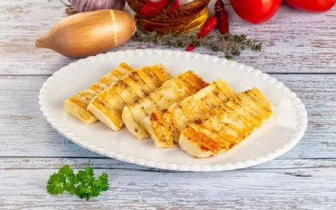 Grillowany ser halloumi na białym talerzu, z dodatkami jak czosnek, cebula i pomidory w tle.