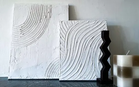 Två vita texturerade tavlor, skapade med spackel, med vågiga mönster. En mörk, taggig skulptur och en rutig ljusbrun och vit ljuslykta står bredvid.