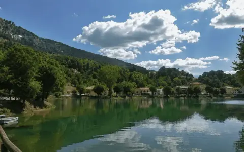 Il lago Sirino, con le sue acque verdi e calme, riflette le case e le colline boscose sotto un cielo azzurro con nuvole bianche.