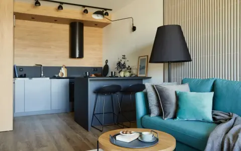 Nowoczesny salon z aneksem kuchennym 22m2. Turkusowa kanapa, czarne hokery przy wyspie, drewniane akcenty i stylowe oświetlenie tworzą przytulną przestrzeń.