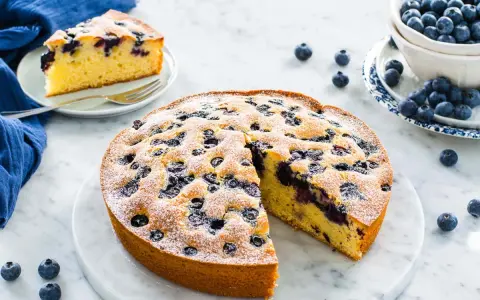 Deliziosa torta allo yogurt con mirtilli, spolverata di zucchero a velo. Una fetta è già servita su un piattino.