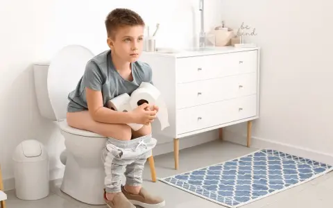 Chłopiec siedzi na toalecie, trzymając rolki papieru toaletowego. Wygląda na chorego, co może sugerować wymioty i biegunka u dziecka.