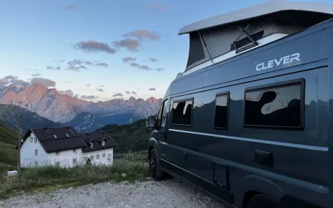 En mörkgrå husbil med pop-up tak, märkt "Clever vans", står parkerad vid en bergig stuga med Alperna i bakgrunden.