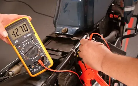 Mit einem Multimeter die Lichtmaschine messen: Ein Mann hält das gelbe Gerät, das -12.70 Volt anzeigt, während die Prüfspitzen an der Motorradverkabelung anliegen.