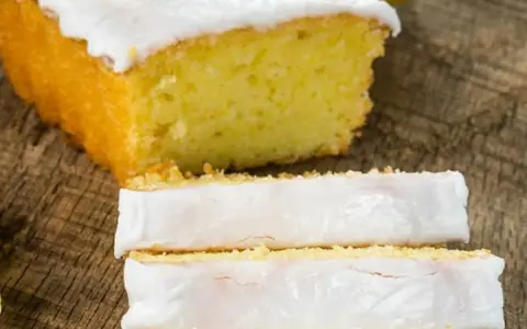 Fetta di plumcake al limone con glassa bianca e una fetta di limone sopra, su un tagliere di legno.