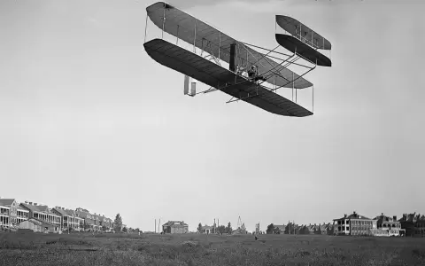 Das Kitty Hawk Flugzeug fliegt über ein Feld mit Gebäuden.