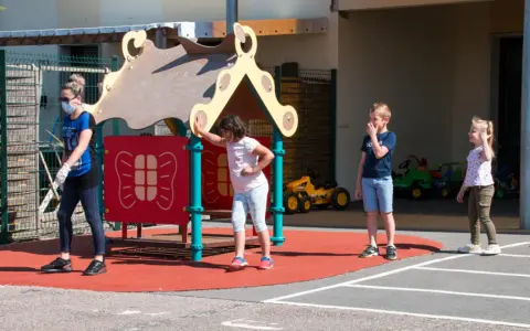 Une animatrice, masque et gants, surveille des enfants sur un terrain de jeu. Le directeur ACM veille à la sécurité.