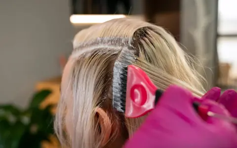 Profesjonalna dekoloryzacja włosów w salonie. Różowa rękawiczka i pędzel nanoszą produkt na blond pasma.