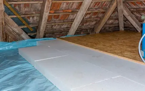 Aislamiento de suelo en una casa antigua con paneles de poliestireno sobre plástico azul, bajo vigas de madera y tejas.