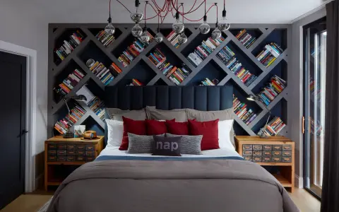 Camera da letto con un'imponente parete di librerie stravaganti a rombi, piene di libri colorati. Un letto accogliente con cuscini rossi e grigi.