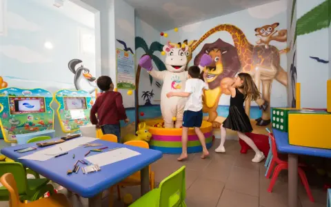 Bambini giocano con un personaggio in costume da ippopotamo in una stanza colorata, un'area giochi per famiglie al Mokambo Shore Easy Family Hotel.