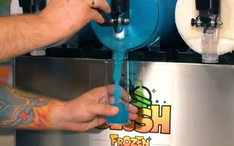Ein Arm mit einem Ketten-Tattoo füllt ein Glas mit blauem Slush Eis aus einem Automaten.