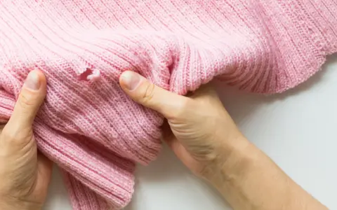 Dłonie trzymają różowy sweter z dziurą, pokazując problem, jak zaszyć dziurę w dresach.