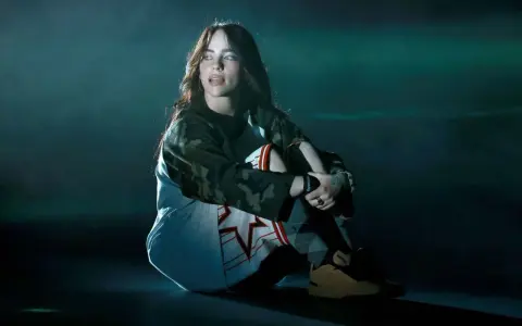 Billie Eilish w ciemnym pomieszczeniu, siedzi w pozie zamyślenia. Jej stylizacja nawiązuje do estetyki jej albumu.