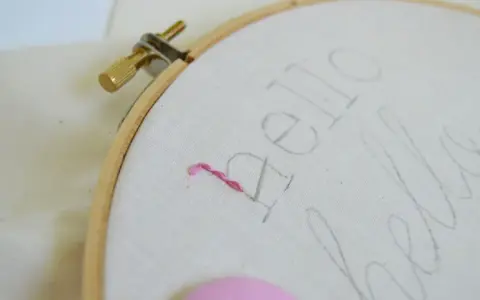 Bastidor con tela y un hilo rosa comenzando a bordar letras.