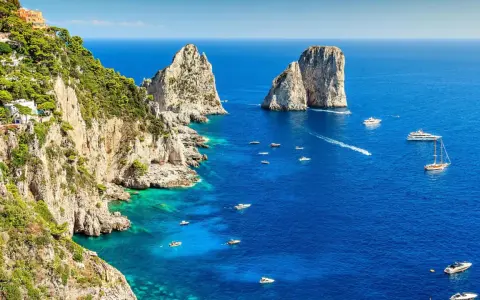 Scogliere verdi e rocciose si affacciano sul blu intenso del golfo di Napoli, punteggiato da barche e yacht. Faraglioni iconici emergono dal mare.